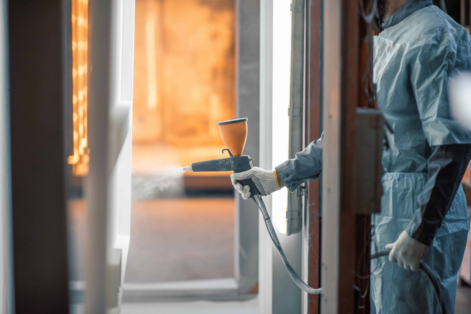 Man wearing a protective suit is powder coating a metal sheet in a spray booth holding a powder gun in his right hand